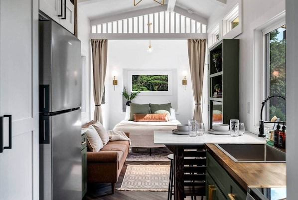 A modern kitchen with a built-in refrigerator, a dining area with a wooden table and chairs, and a cozy bedroom with a bed and a view of greenery.