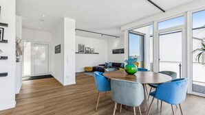 A modern living room with a wooden floor, a round dining table, and blue chairs. There is a sofa and a yellow armchair in the background.