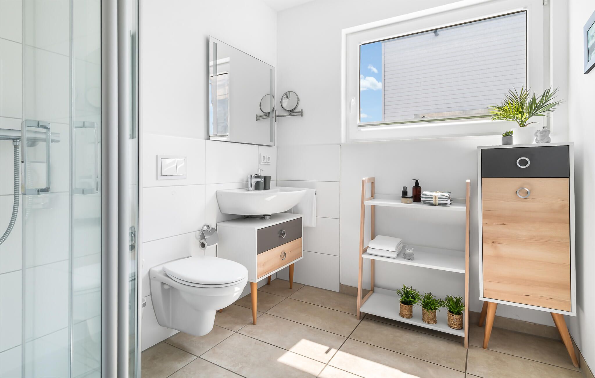 Une salle de bain moderne avec une douche à l’italienne en verre, des toilettes blanches, un lavabo avec un miroir au-dessus et un meuble-lavabo en bois doté d’étagères.