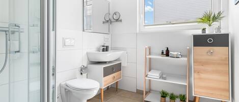Une salle de bain moderne avec une douche à l’italienne en verre, des toilettes blanches, un lavabo avec un miroir au-dessus et un meuble-lavabo en bois doté d’étagères.