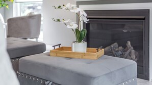 Una sala de estar moderna con un sofá gris, una bandeja de madera con un jarrón blanco y una orquídea, y una chimenea al fondo.
