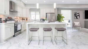 Una cocina moderna con armarios blancos, una isla central con taburetes de bar, electrodomésticos de acero inoxidable e iluminación colgante.