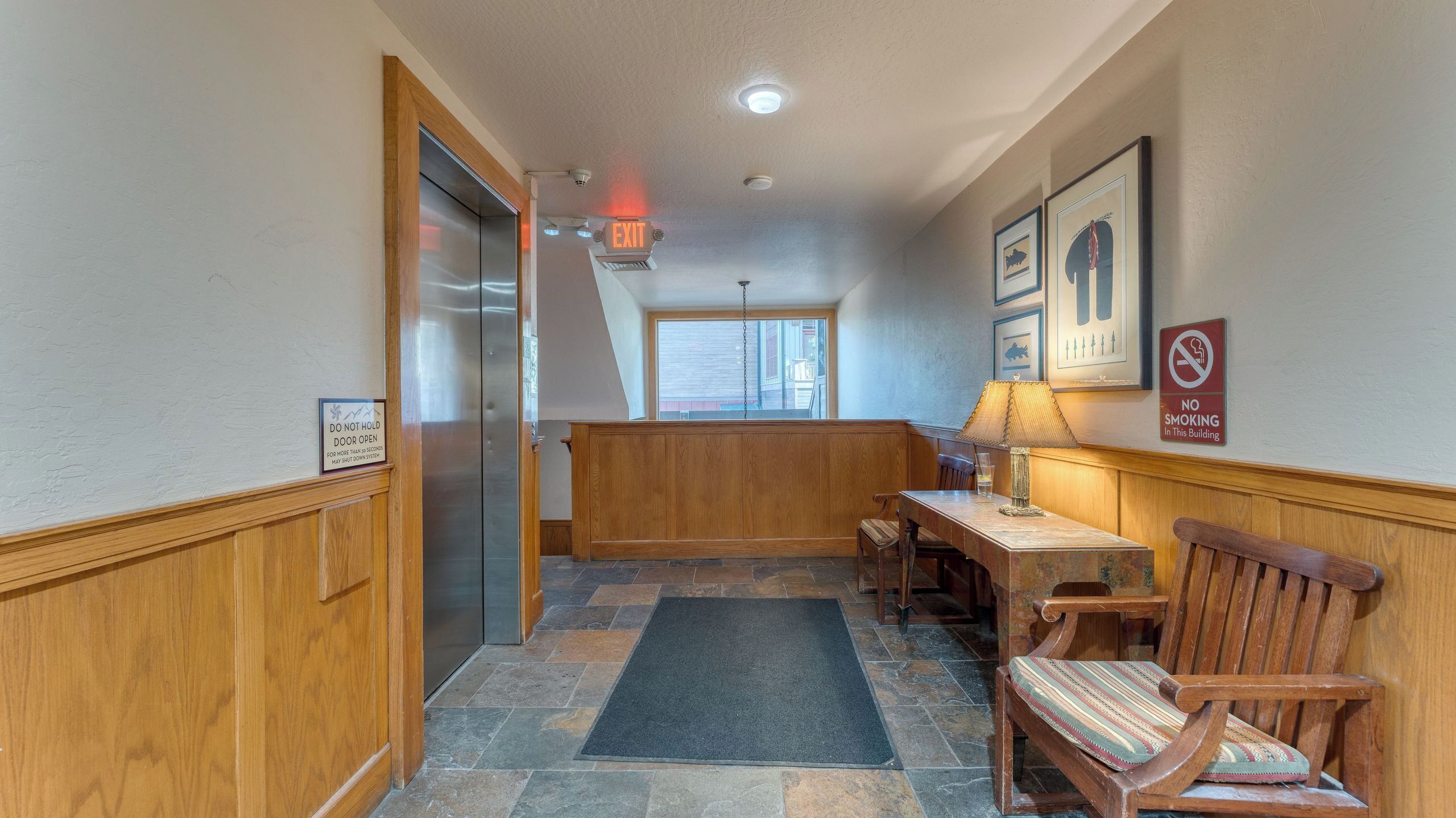 En hotels korridor med en træbænk, et bord med en lampe, en dør med et skilt der står 'DO NOT HOLD DOOR OPEN', og et 'RYGNING FORBUDT' skilt på væggen.