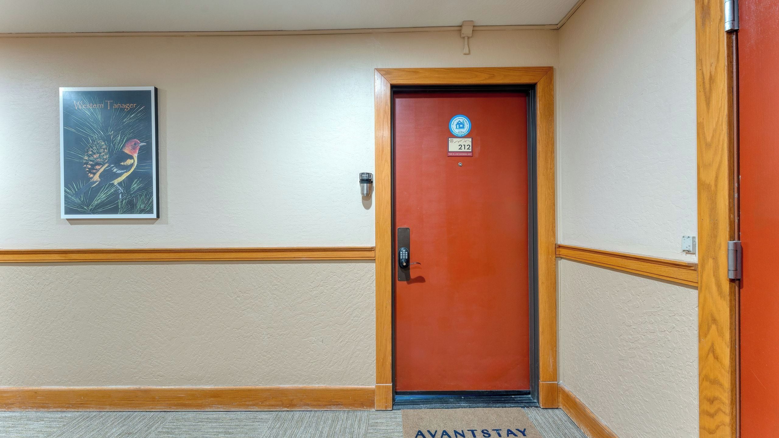 A hotel entrance with a red door, a security keypad, a doormat, and a framed poster on the wall.