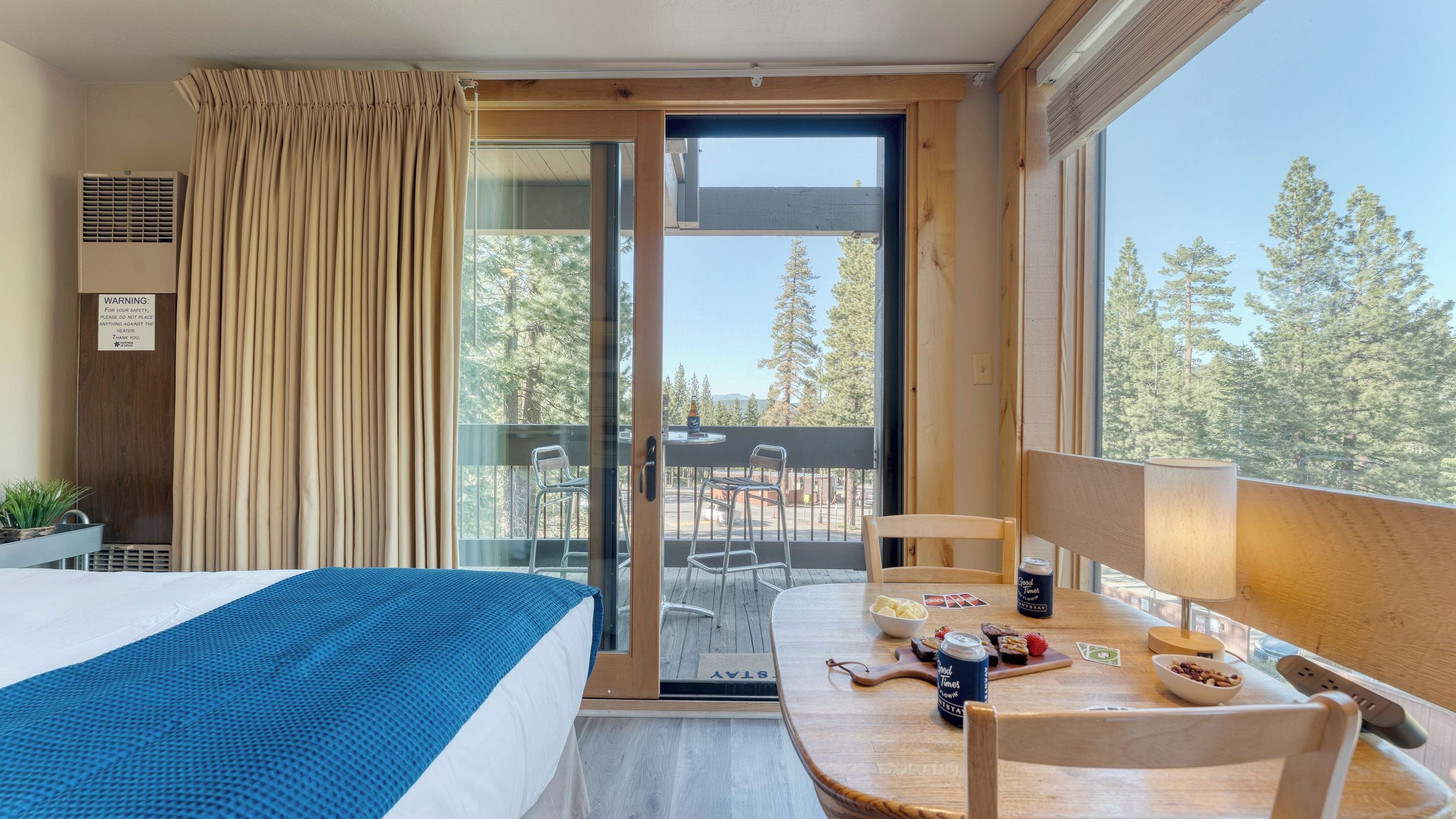Una camera d'albergo con un letto, un tavolo con degli snack, una sedia, un balcone che si affaccia su alberi e un cartello che avverte sui rischi del termosifone.