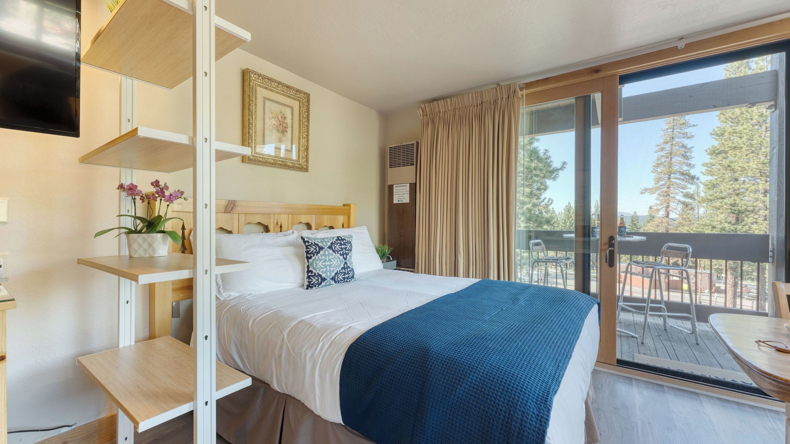 A hotel room with a bed, a desk, a TV, a window with curtains, and a balcony with outdoor furniture.