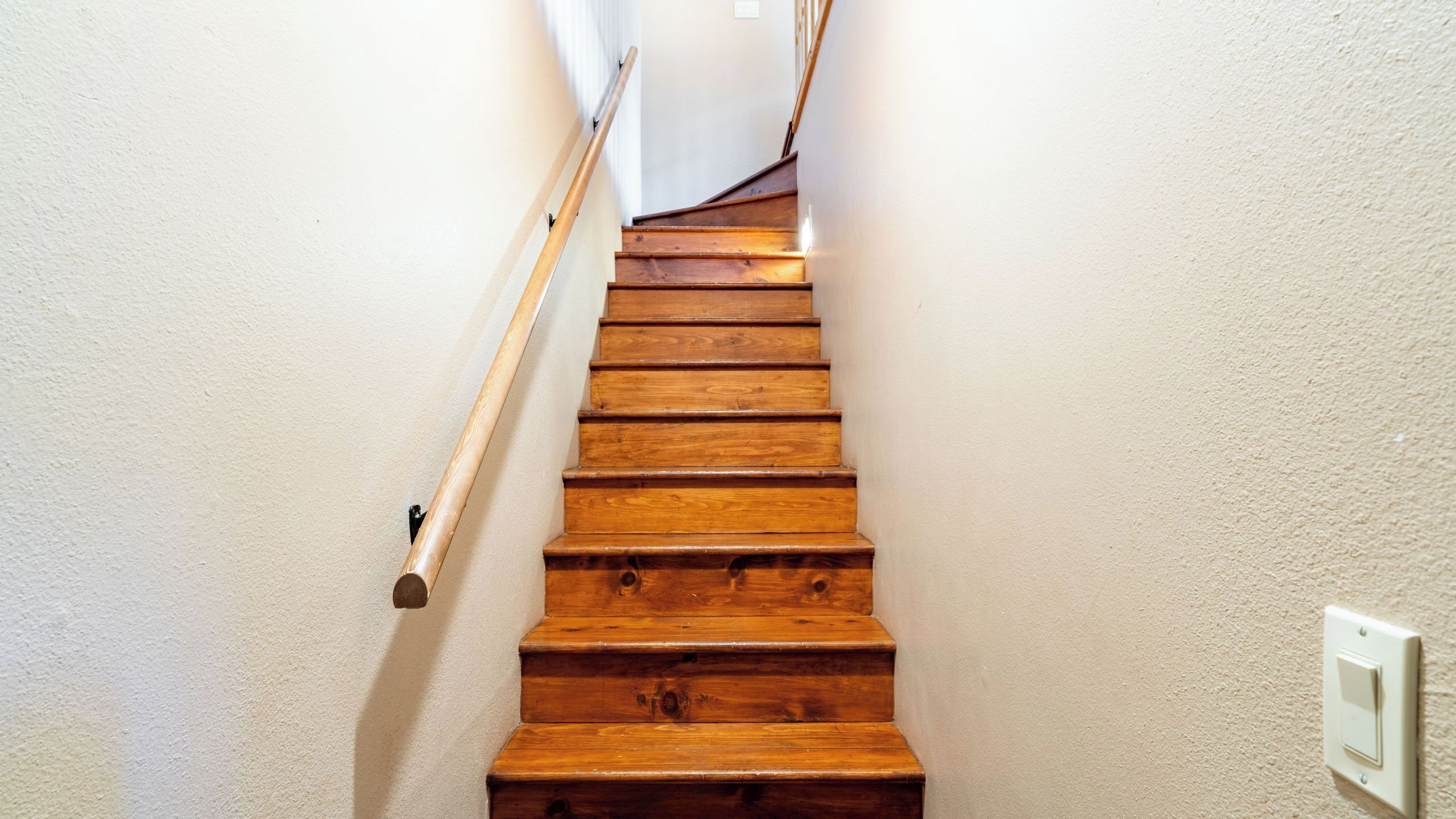 Una escalera de madera con barandilla, paredes blancas y un interruptor de luz en la pared.