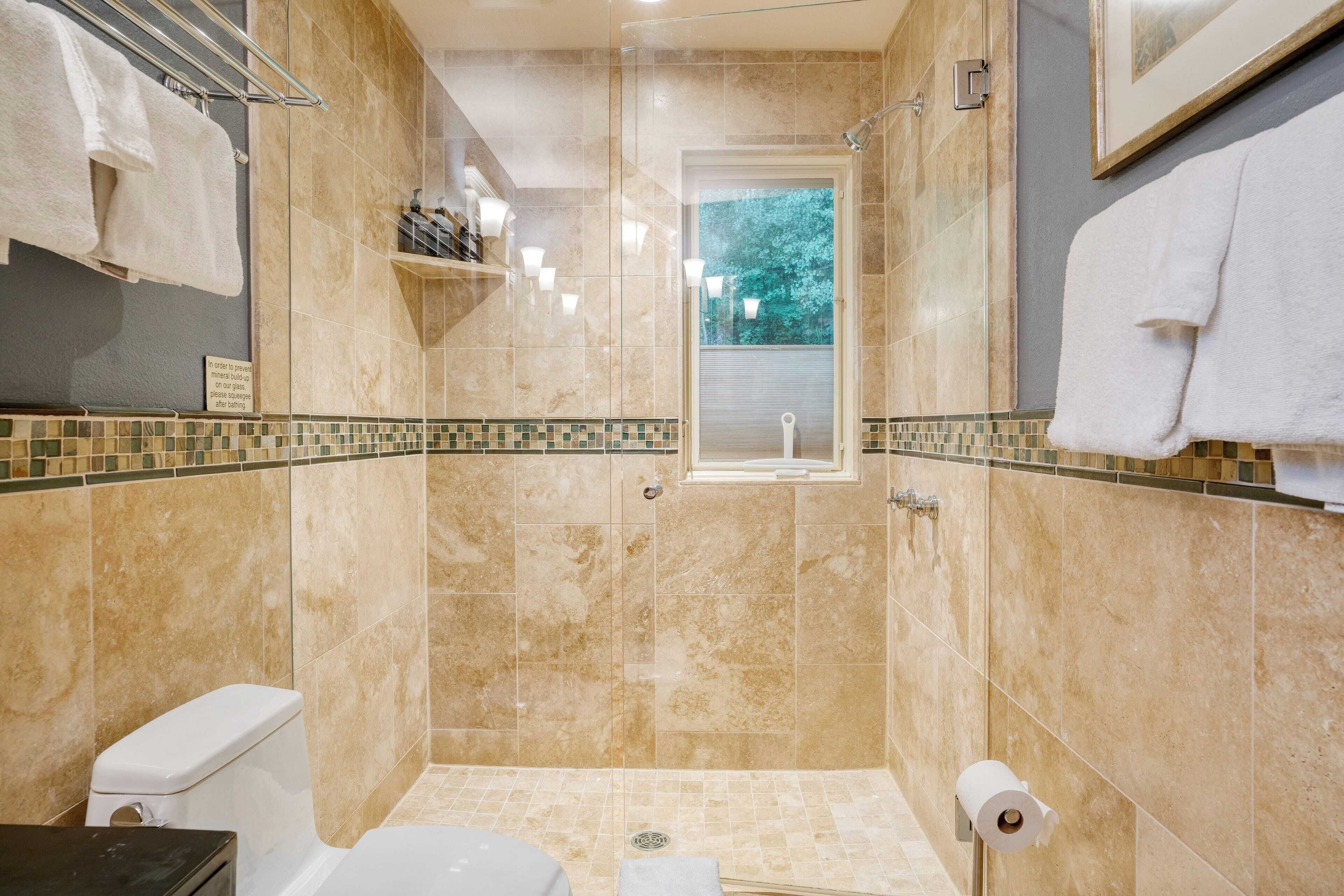 A bathroom with a shower, toilet, and sink, featuring beige tiles and a small window.