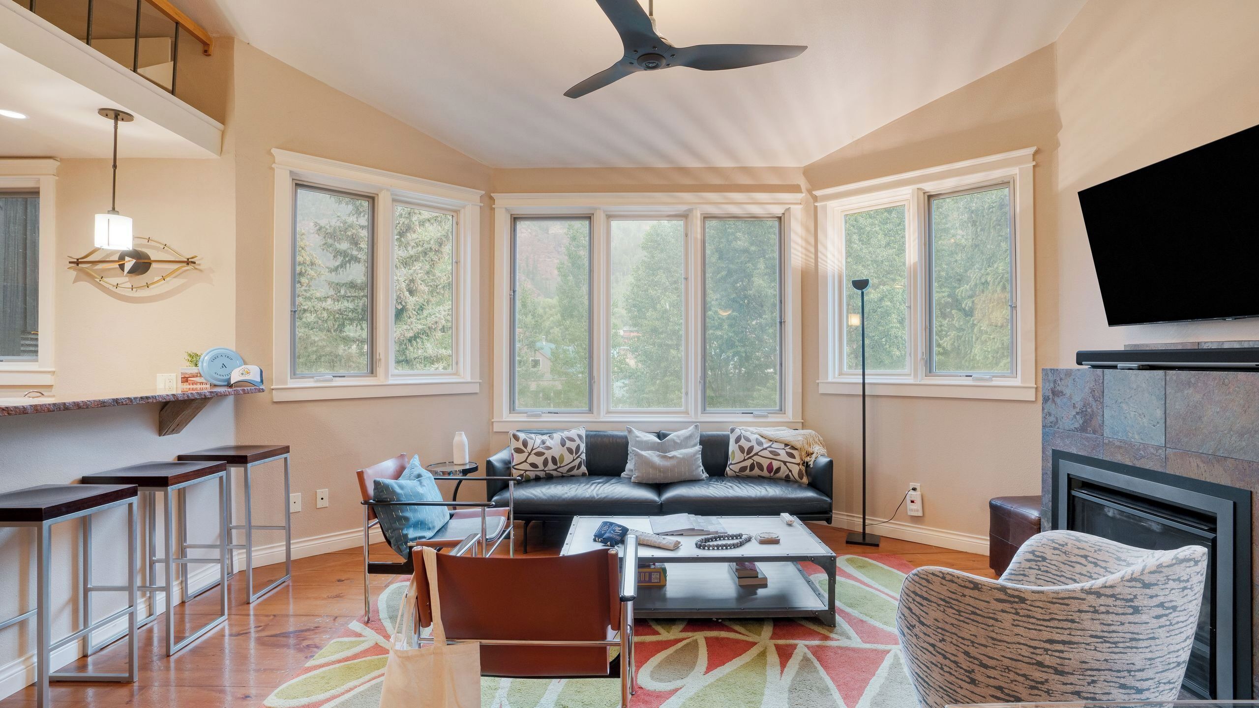 A spacious living room with a high ceiling fan, large windows, a fireplace, and a modern kitchen area.