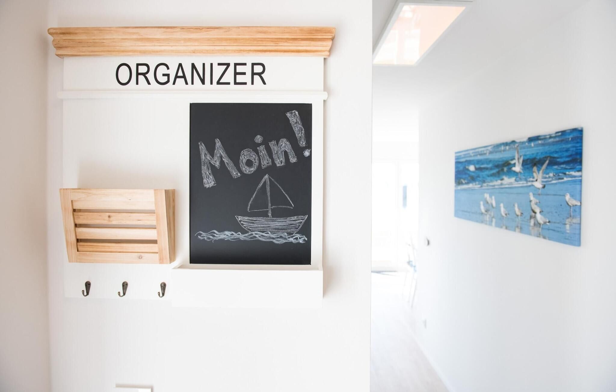 Un mur avec un tableau noir sur lequel on peut lire « Moin ! » et un petit dessin de bateau, un organiseur en bois et un tableau représentant un goéland.