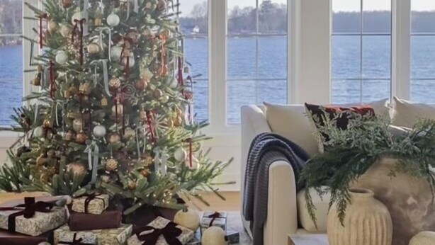 Uma árvore de Natal decorada com presentes em um quarto com janelas amplas com vista para um corpo de água.