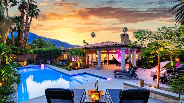 A poolside area with lounge chairs, a table with a bottle and candles, and a building with a tiled roof and columns. - Amazing- Ultra Luxe Estate, Resort like Pool, Firepit & More (Palm Springs)