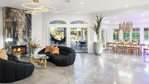 A spacious living room with a fireplace, black sofas, a glass coffee table, and a dining area with wooden chairs. - Amazing- Ultra Luxe Estate, Resort like Pool, Firepit & More (Palm Springs)