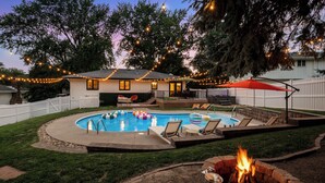 A backyard with a swimming pool, lounge chairs, a fire pit, and string lights. - Omaha Oasis! Pool, Hot Tub, Mini Golf, Bar, Games (Omaha)