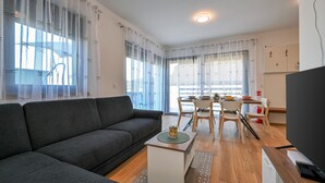A modern living room with a grey sofa, a dining area with a table and chairs, and a TV on a wooden stand. - 2 bedroom cozy home in Margecan (Margecan)