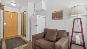 A compact living space with a brown sofa, a white refrigerator, a wooden door, and a painting of a forest. - Comfortable Studio in the Heart of Park City (Park City)