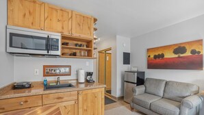 A compact kitchen with wooden cabinets, a microwave, and a coffee maker. There is a gray sofa and a painting of trees on the wall. - Comfortable Studio in Park City 325 (Park City)