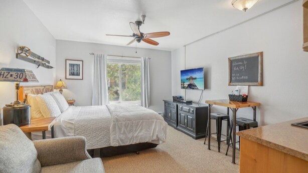 A bedroom with a bed, a desk, a chair, a ceiling fan, a window with curtains, a TV, and a welcome sign. - Comfortable Studio in Park City 325 (Park City)