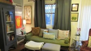 A living room with a green sofa, a small table, a bookshelf, and a window with curtains. - Private Detached Cottage ~Near Downtown Phoenix! (Phoenix)