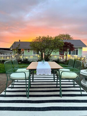 A dining area with a table set for four, surrounded by green chairs with patterned cushions, under a sunset sky. - Camano Island Cottage - Madrona Beach (Camano Island)
