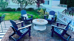 Eine Terrasse mit Adirondack-Stühlen und einem Feuerkorb, umgeben von einem weißen Zaun und Blick auf einen See.