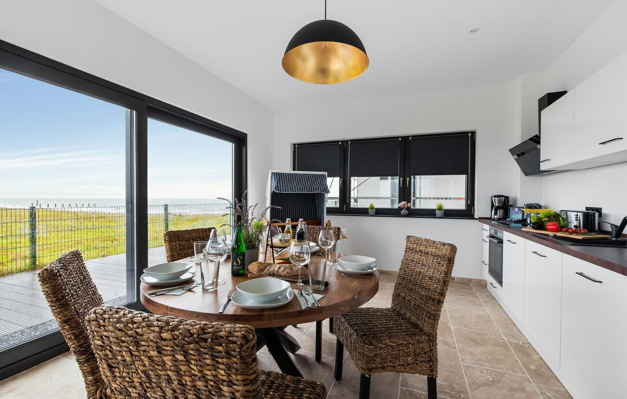 A modern kitchen with a large dining table, wicker chairs, and a view of the ocean.