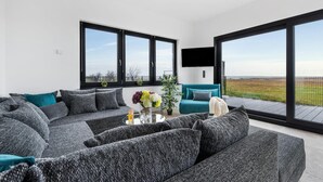 Ein modernes Wohnzimmer mit einem Ecksofa, einem Türkis-Armlehnstuhl und einem Couchtisch mit Blumen. Große Fenster bieten Blick ins Grüne.