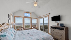 A bedroom with a large bed, a ceiling fan, a dresser, a television, and a view of the ocean through large windows. - 7865- Peridot, Oceanfront 5 bedroom home in Frisco! (Frisco)