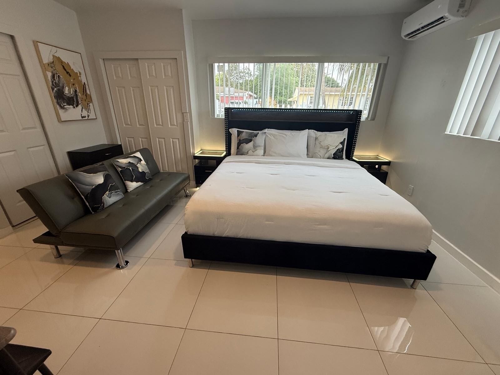 A bedroom with a large bed, a bench, a bedside table, and a window with blinds.