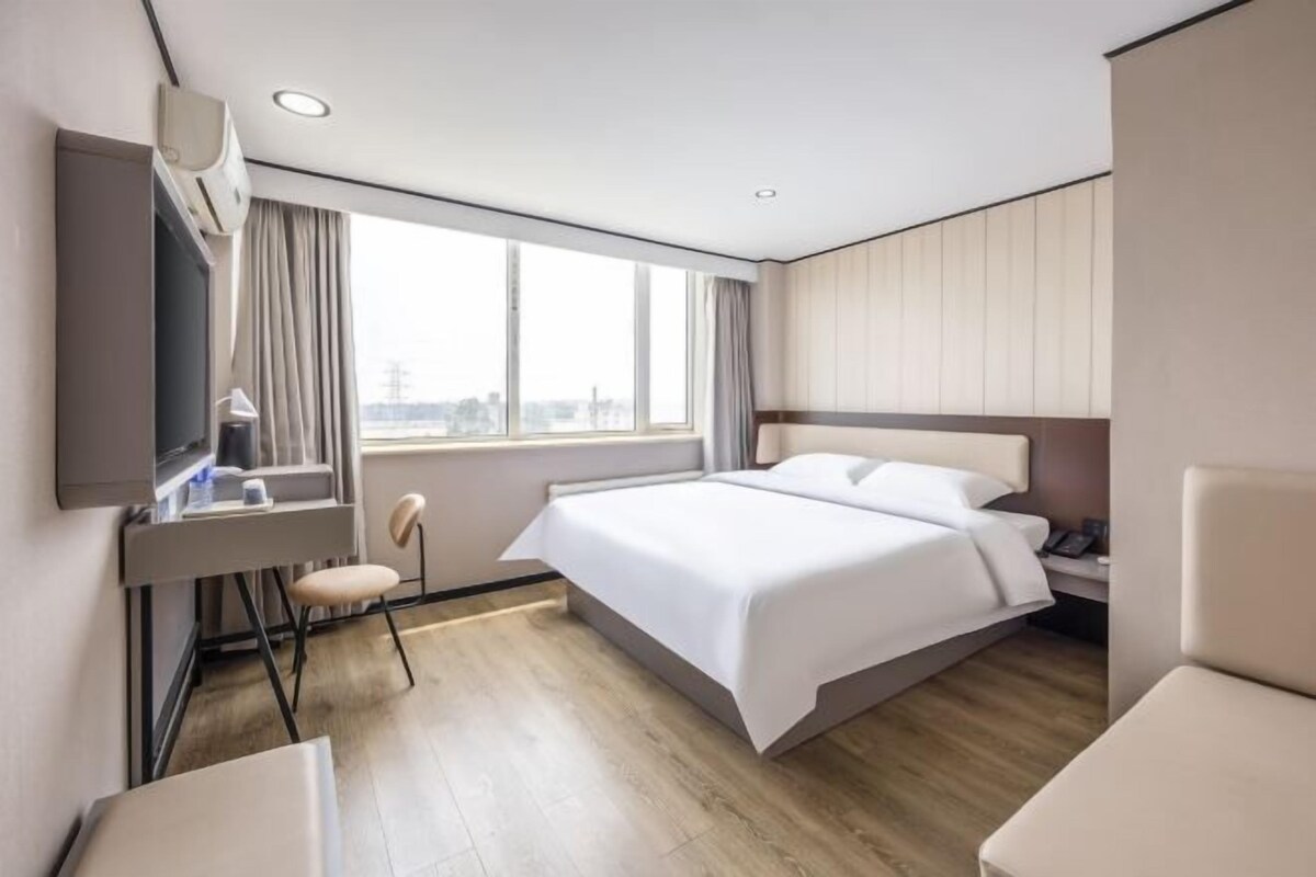 A modern hotel room with a large bed, a desk with a chair, a TV, and a window with a city view.