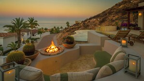 A cozy outdoor seating area with a fire pit, cushions, and a view of the ocean.