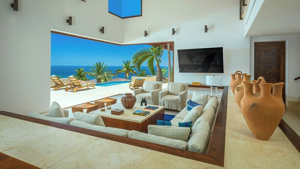 A spacious living room with a large window offering a view of the beach and ocean. There is a comfortable seating area with a sofa and chairs, a coffee table, and a large vase.