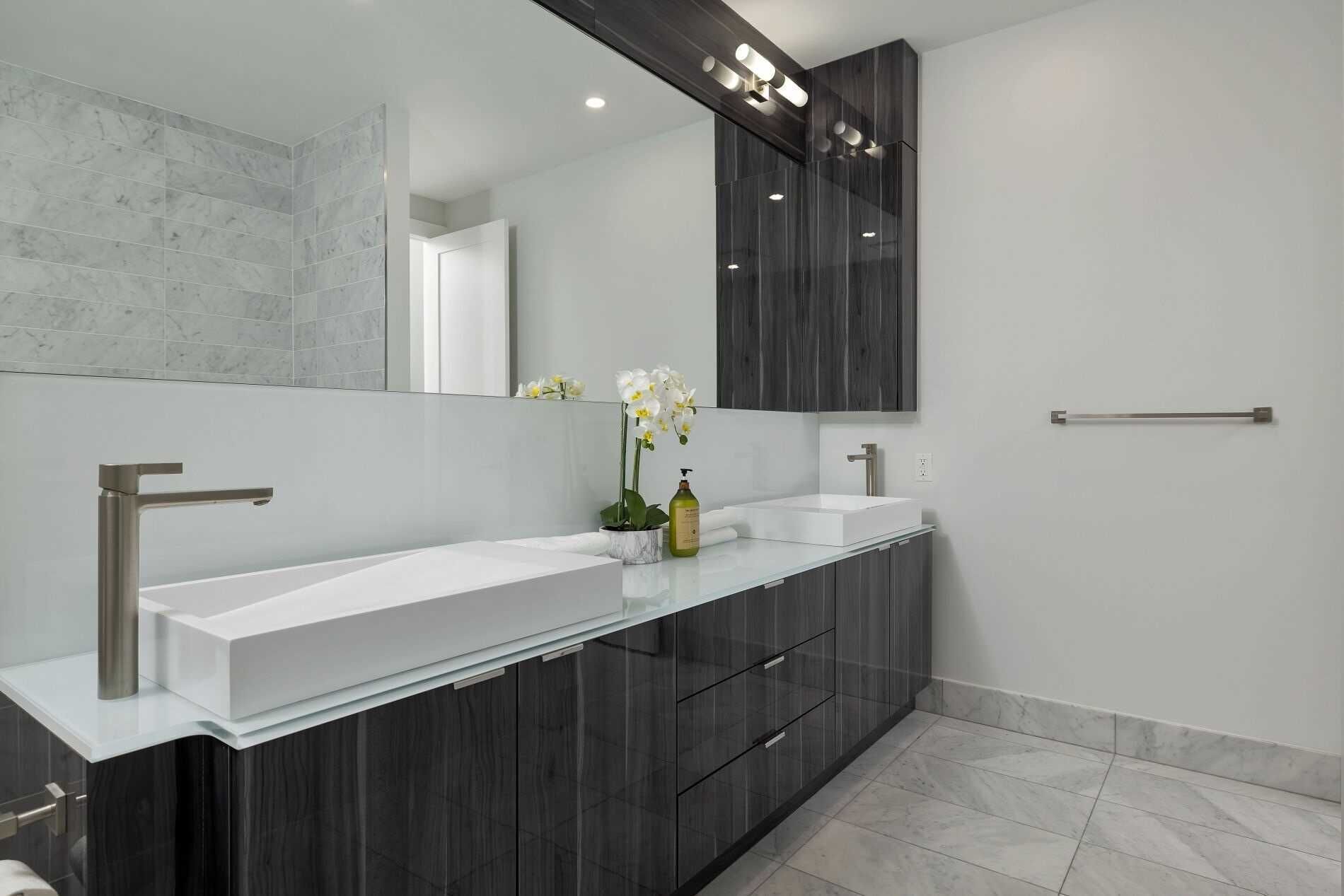 A modern bathroom with a large mirror, two sinks, dark cabinetry, and a glass countertop.