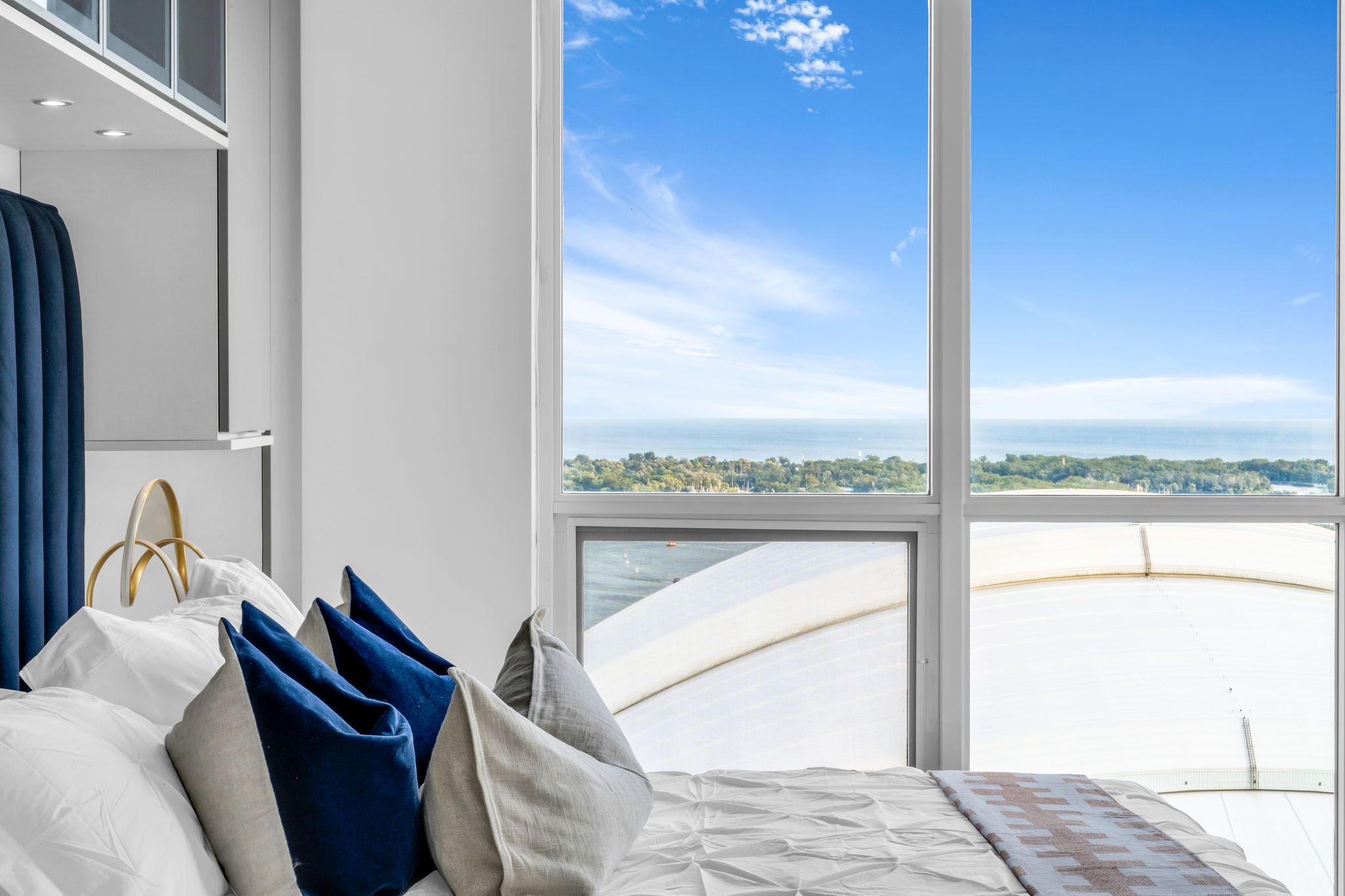 A bedroom with a large window, a bed with pillows, and a view of a beach and cityscape.