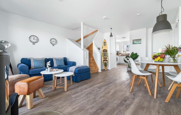 A modern living room with a blue sofa, wooden coffee table, and a dining area with white chairs and a wooden table.