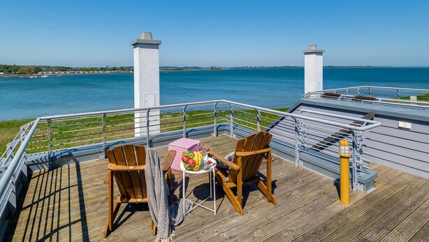 Une terrasse avec deux chaises en bois et une petite table, donnant sur une étendue d’eau dont on aperçoit la rive au loin.