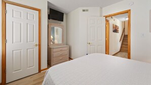 Une chambre à coucher avec un lit, une commode et un miroir. Une porte donne sur un couloir où se trouve un escalier.