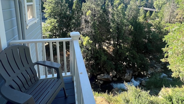 A deck with a chair, overlooking a forested area with a stream and a mountain in the background.