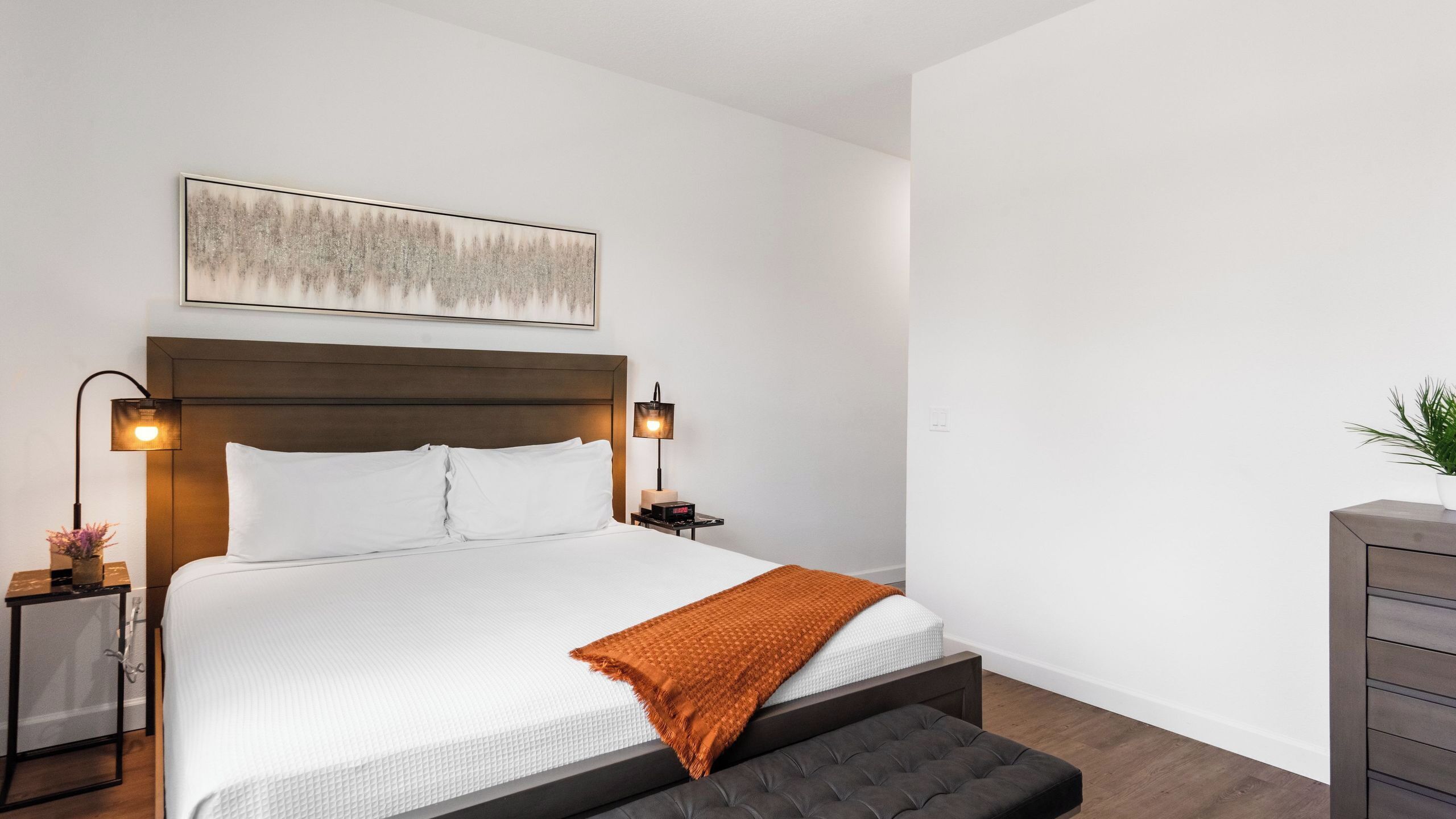 Un lit bien fait, avec une couette blanche et un plaid marron, deux lampes de chevet, une tête de lit et une œuvre d’art accrochée au mur.