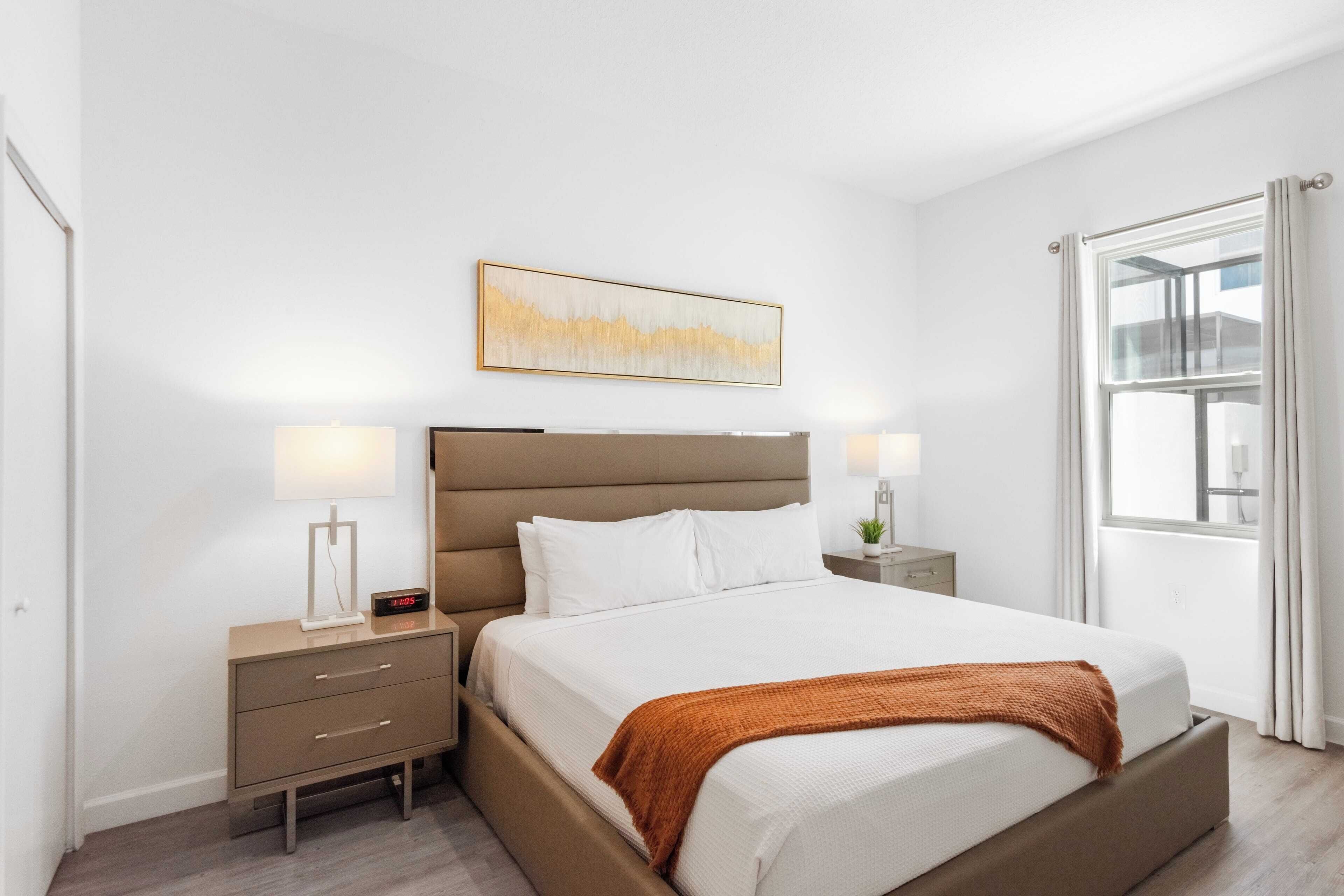 A modern bedroom with a large bed, two bedside tables with lamps, a nightstand, a window with curtains, and a wall-mounted abstract painting.