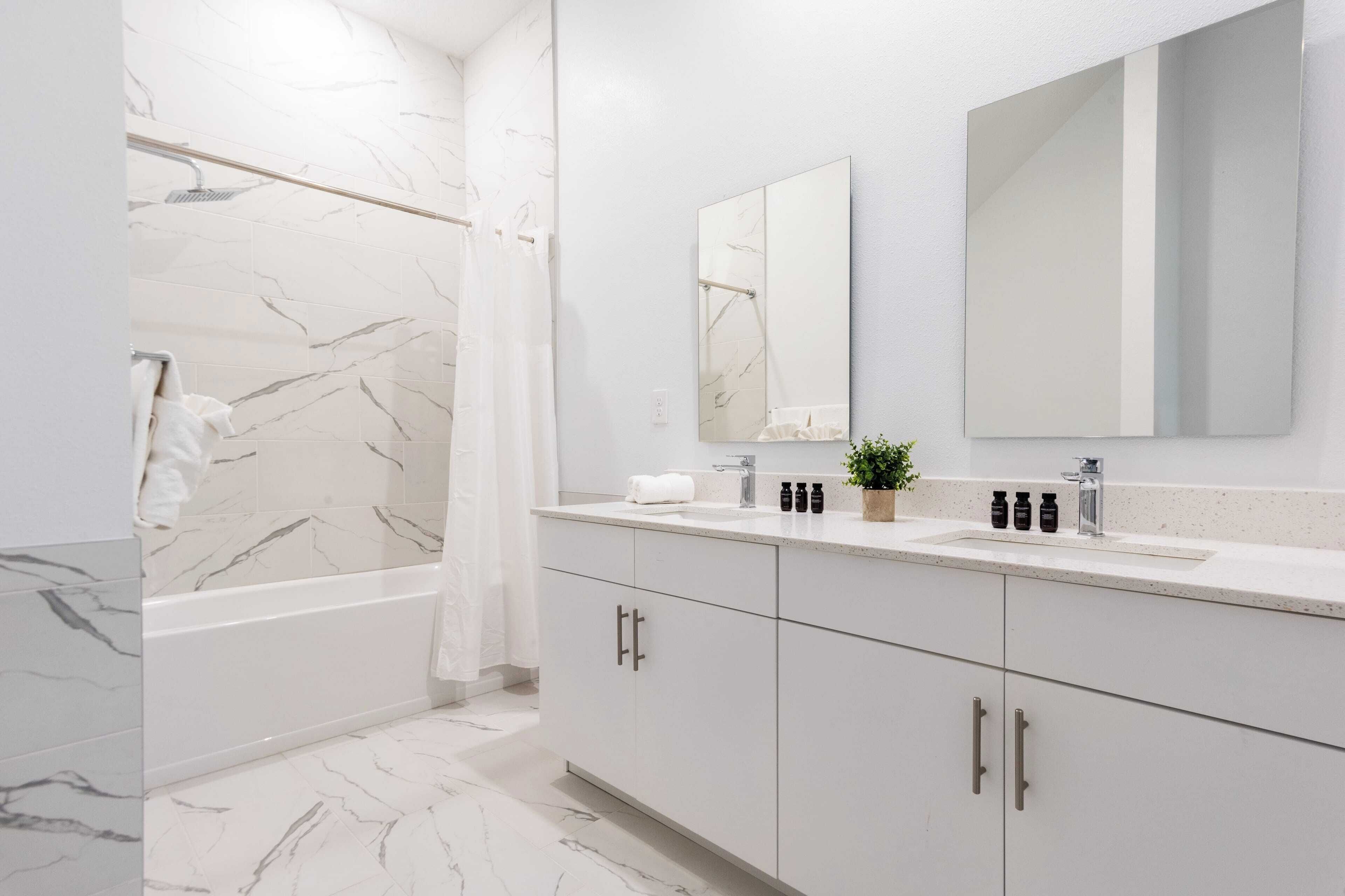Une salle de bain moderne avec un sol en marbre, une baignoire, un lavabo avec un miroir au-dessus et une plante en pot posée sur le comptoir.