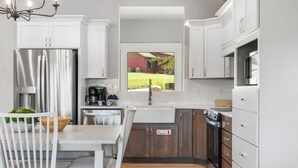 A modern kitchen with white cabinets, stainless steel appliances, a dining area with a white table and chairs, and a view of a red house through a window. - New Home in Trophy Run with Private Hot Tub & Firepit, Near Taneycomo & Table Rock, Free Tickets (Branson)