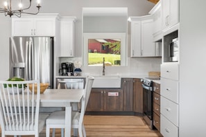 Eine moderne Küche mit weißen Schränken, Edelstahlgeräten, einem Essbereich mit weißem Tisch und Stühlen sowie Blick auf ein rotes Haus durch ein Fenster.