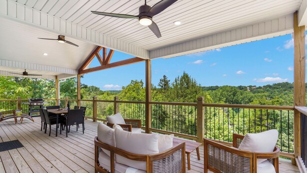 A covered outdoor deck with wooden furniture, a grill, and a view of a forested landscape. - New Home in Trophy Run with Private Hot Tub & Firepit, Near Taneycomo & Table Rock, Free Tickets (Branson)