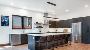 A modern kitchen with a central island, bar stools, stainless steel appliances, and a large window. - New! Home Again Luxury Haven - Sleeps 8 (Whitefish)