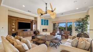 A spacious living room with a large TV, comfortable seating, a fireplace, and a ceiling fan. - Villa Paradiso - 2 Pools, 5 Suites & More! (Indio)