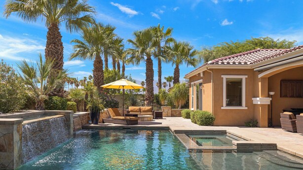 A pool area with clear water, surrounded by palm trees and a building with a tiled roof. - Villa Paradiso - 2 Pools, 5 Suites & More! (Indio)
