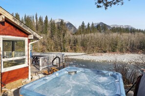 Une cabane en bois dotée d’un bain à remous et d’une terrasse offrant une vue sur une rivière et des montagnes.