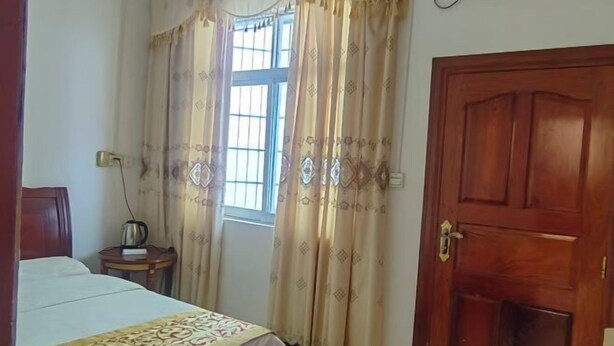 A bedroom with a bed, a bedside table with a kettle, a window with curtains, and a wooden door. - Dongxing Wanwei Jintan Xiangyue Hotel (Dongxing)