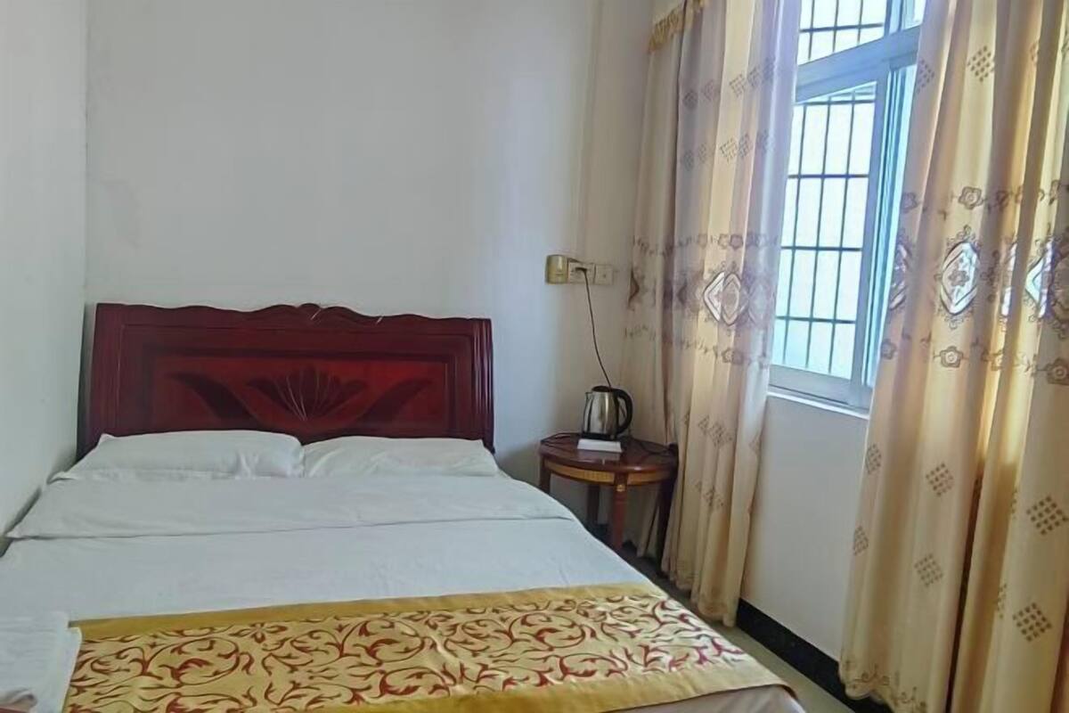 A hotel room with a wooden headboard, a bed with white linens, a patterned throw, a small bedside table with a kettle, and a window with patterned curtains.